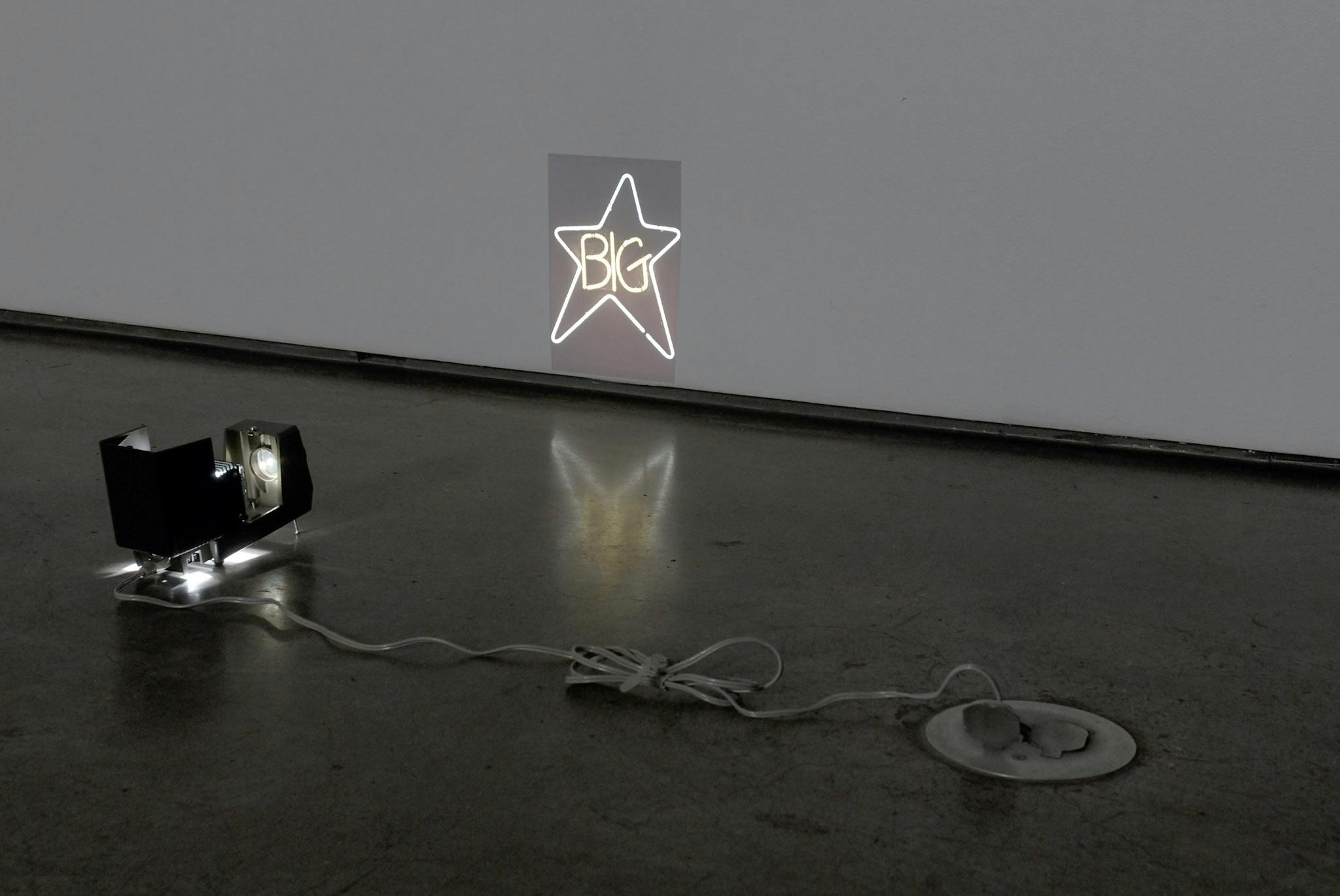 A small light is placed on the gallery floor. The shape of this light resembles an old accordion camera. It projects a shape of a star, with the word BIG written inside, on the wall.