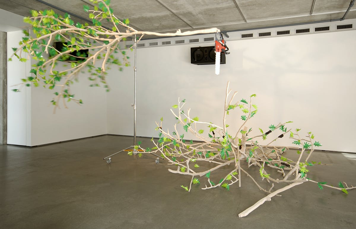 Two large tree branches are in the gallery. One branch hangs from the ceiling and the other sits on the ground. The branch on the ceiling has a chainsaw with a red handle hanging off it. 