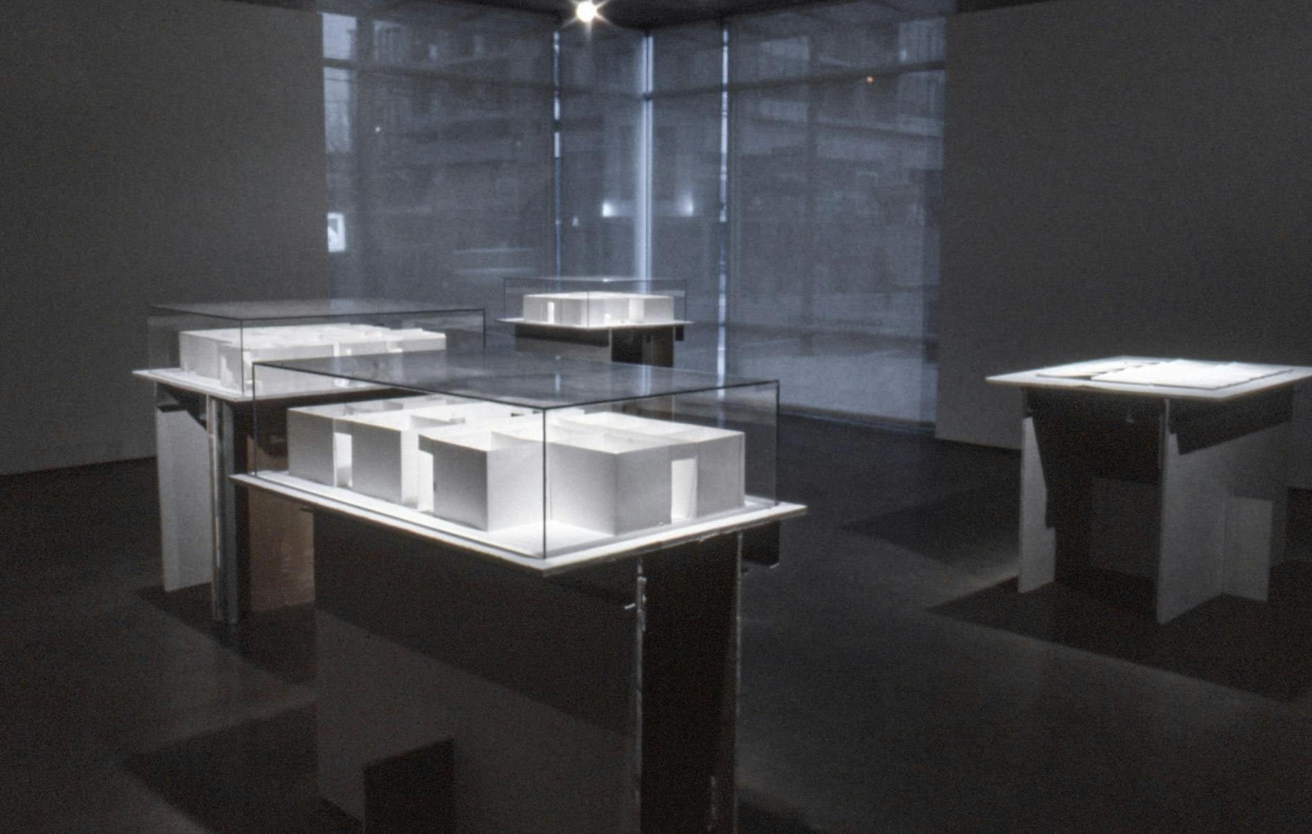 White sculptures are installed in a gallery space. Each sculpture displays a mockup of an architecture complex. They are placed on pedestals made of the cutout parts of the gallery wall. 