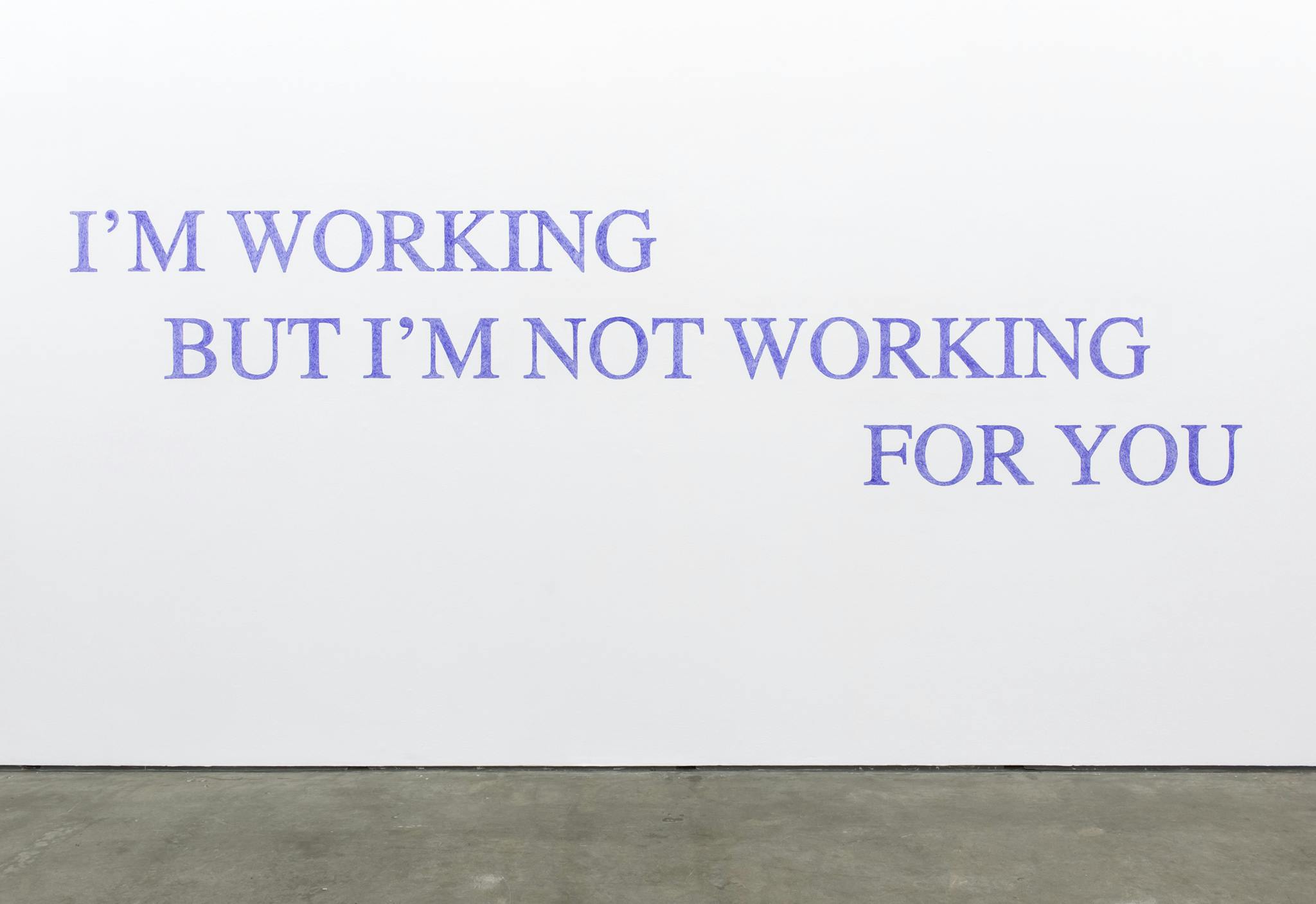 Large blue text on the wall of a gallery space that reads “I’M WORKING BUT I’M NOT WORKING FOR YOU.”