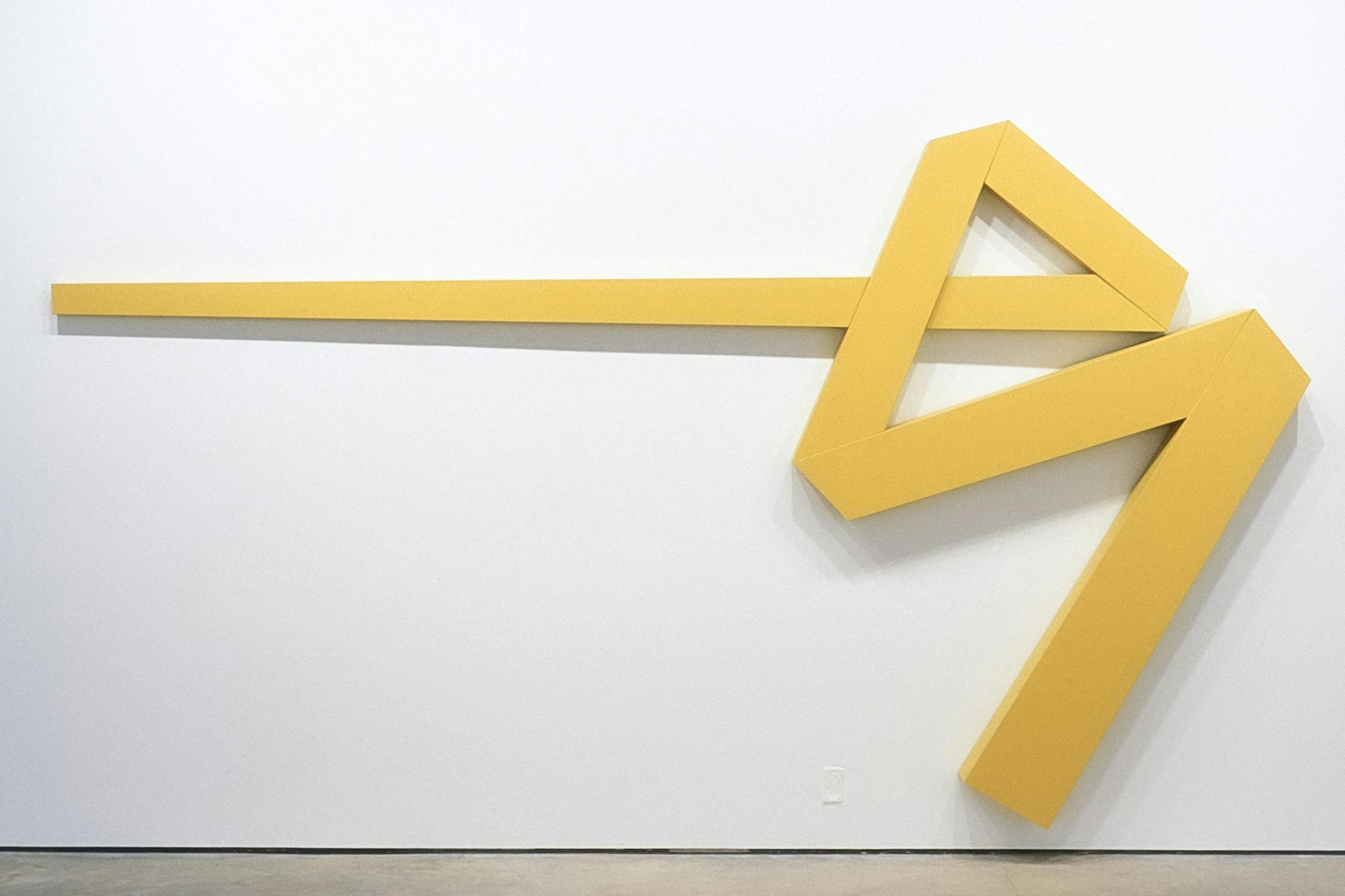 A large-sized geometric sculpture is installed on the gallery wall. Five yellow lines form a shape that resembles an arrow with an extra line that extends downwardly from the arrow point.