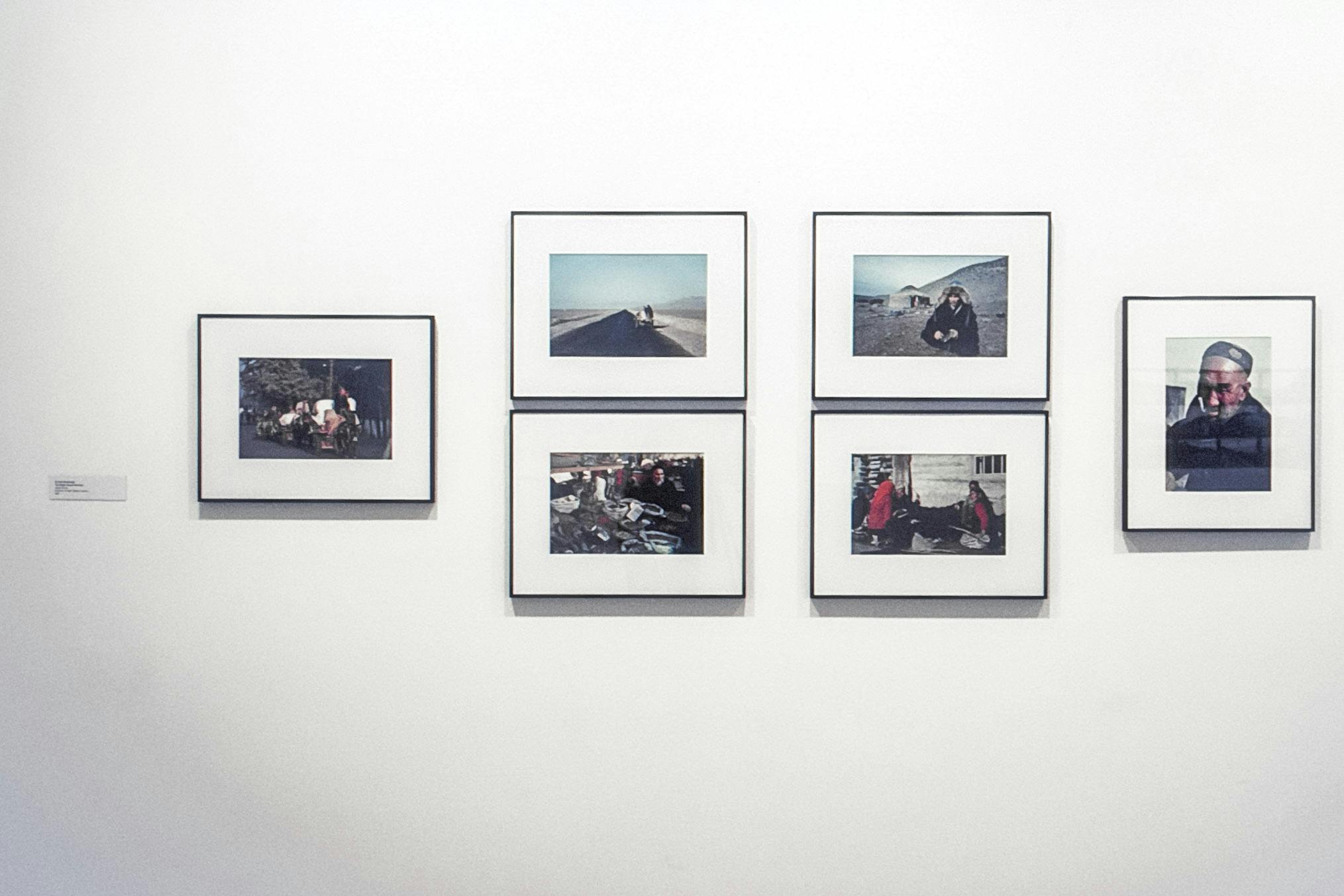 Six photos in black frames displayed on a white wall. The photos show people in different spaces doing a variety of things - some are walking down a stretch of highway, some are selling vessels.
