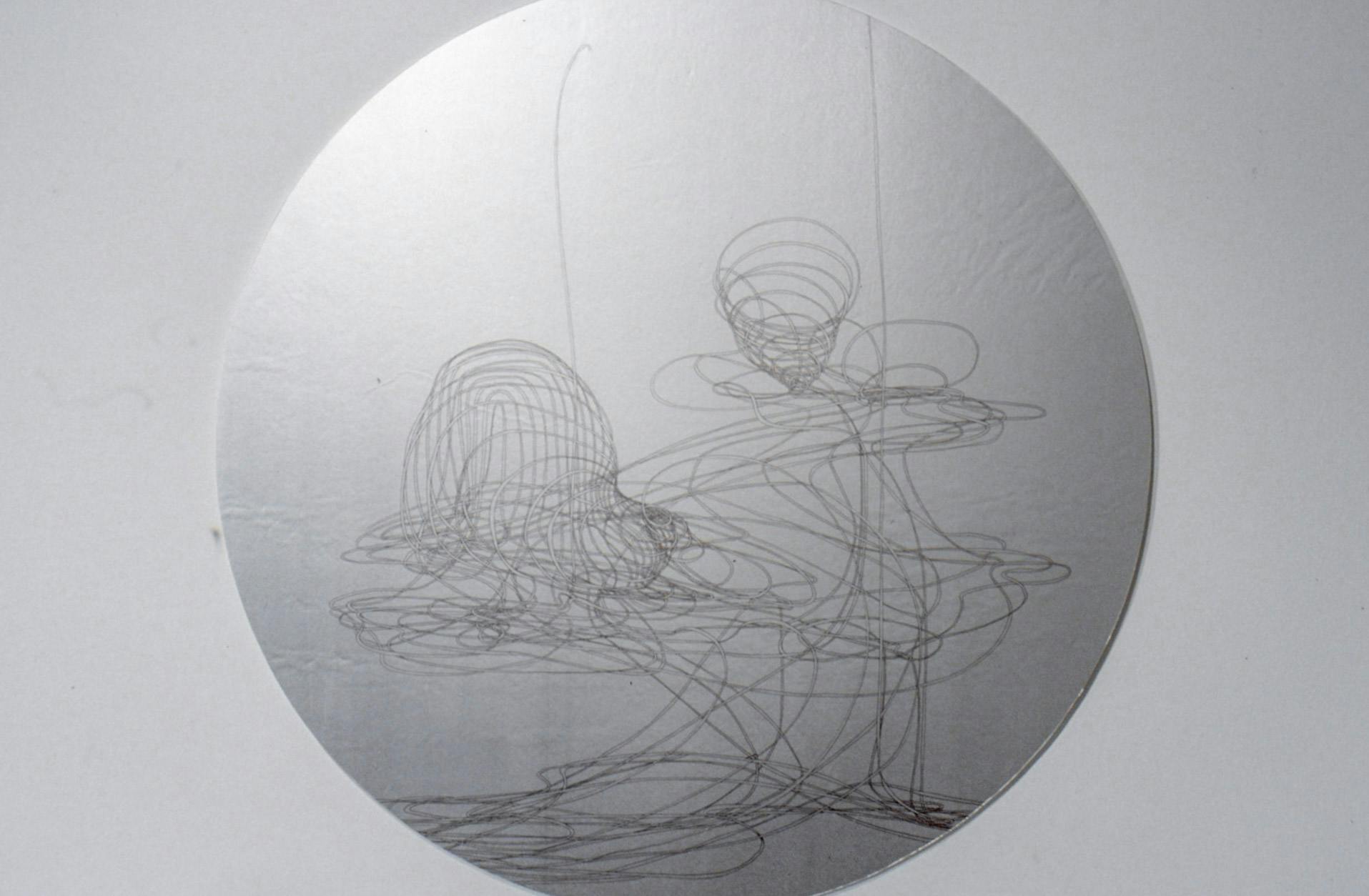 This is a close-up view of a circular shaped sculpture made by Lucy Pullen. On its silver metallic surface, an abstract line drawing is made. The drawing looks like two marionettes dancing together. 