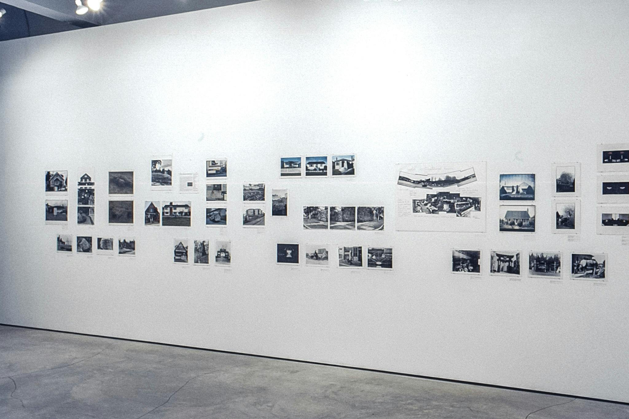 A white wall in a gallery with dozens of different photos. Some are colour, some are black and white. The majority of the photos show the exterior of several different buildings. 