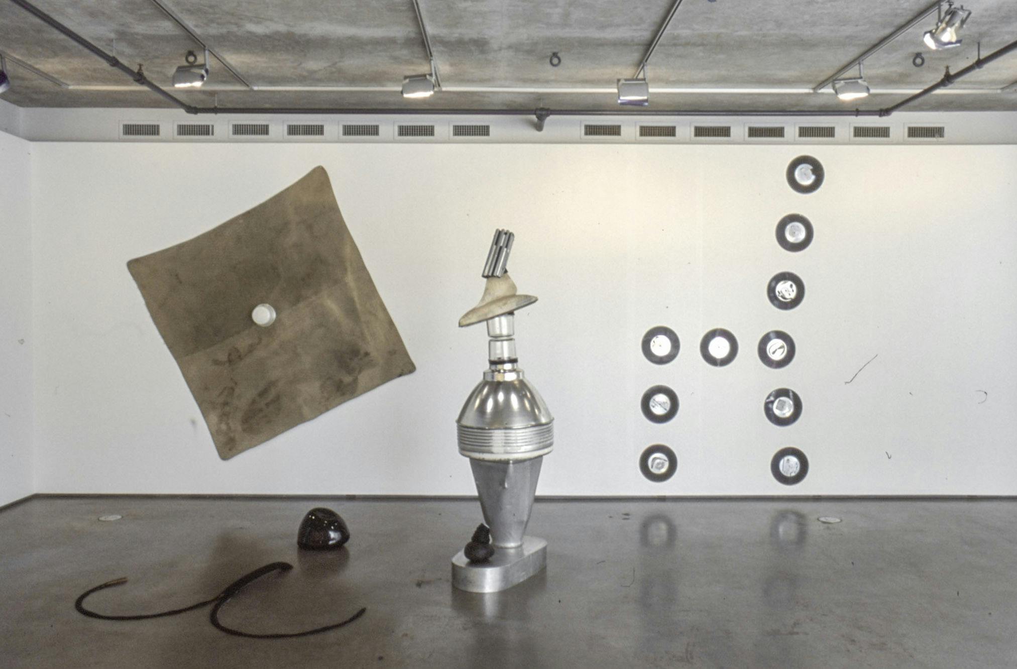 Installation image of Jerry Pethick’s artworks. A black round object and a lope-like object are on the floor next to a tall machine-like sculpture. A large square piece of brown fabric and 10 silver circles are mounted on the wall. 