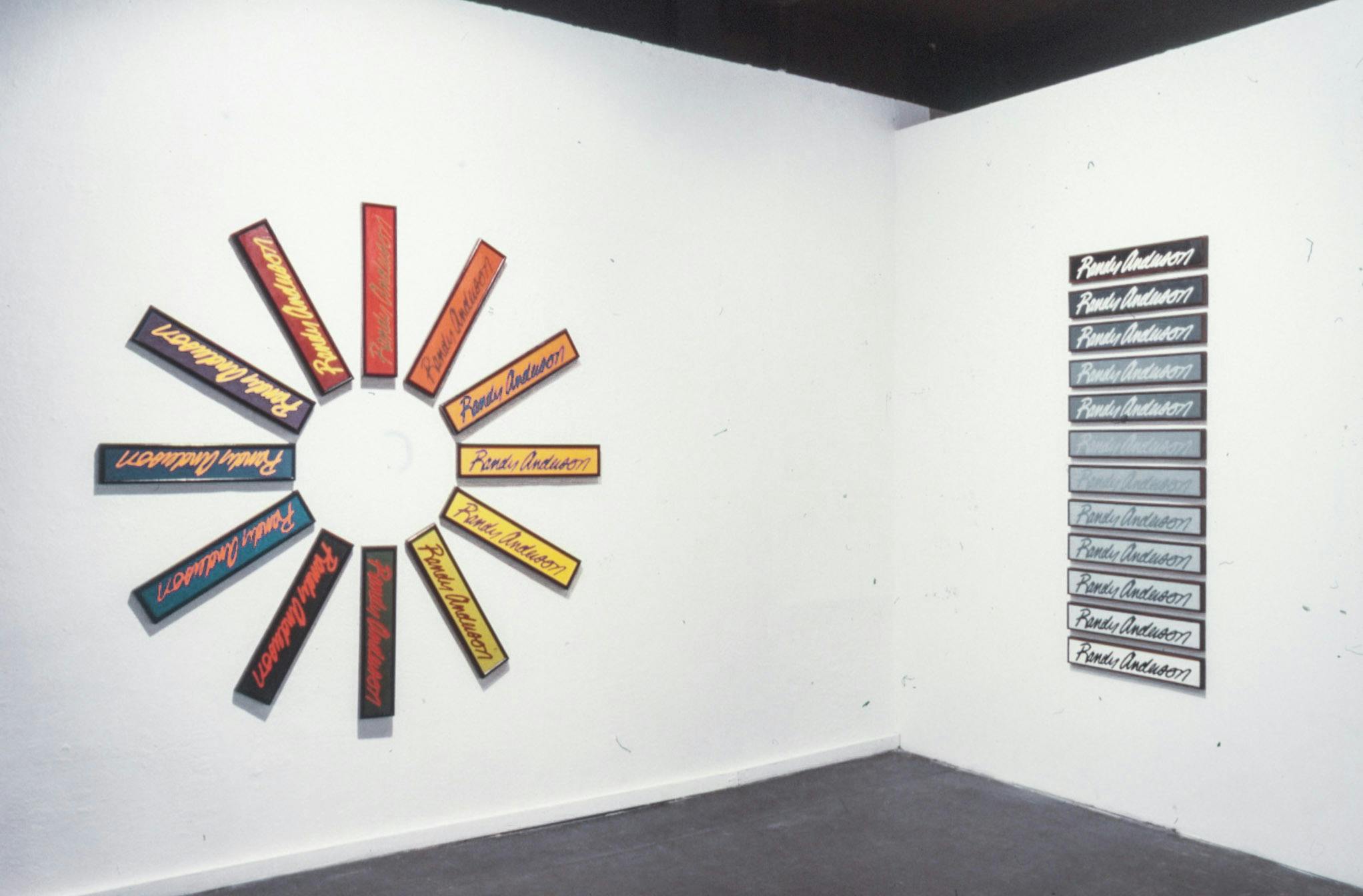 Two artworks made of plaques reading "Randy Anderson” hang on a gallery wall. One  is arranged in a wheel with colourful plaques, the other has plaques in a column going from black to white.