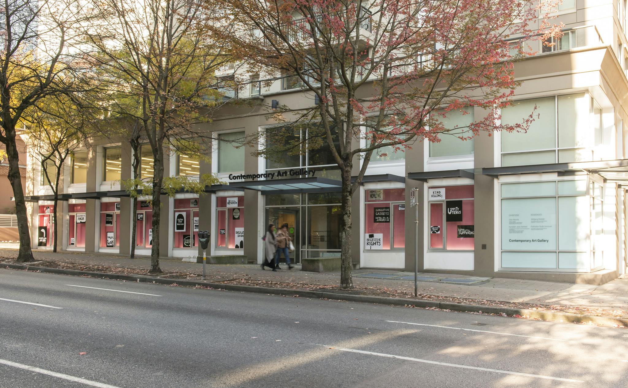 An exterior image of CAG’s façade with the work of Kameelah Janan Rasheed installed on the windows. Each window has a pink backdrop with black and white text-based vinyl prints scattered across. 