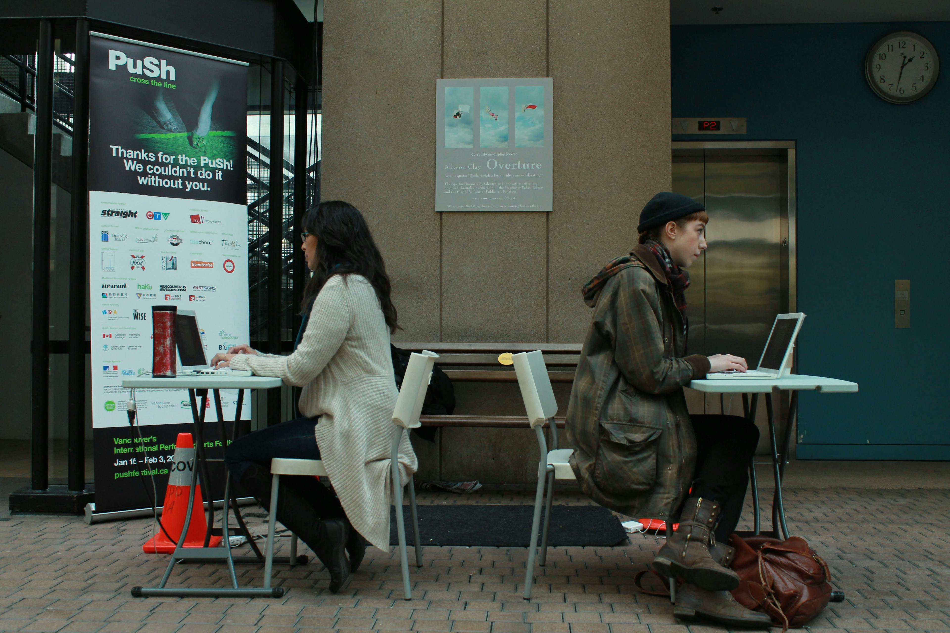 Two people sit back-to-back at folding tables in a public space. Both are typing on white laptops. A large poster for Push Festival is visible behind one desk, an elevator door behind the other.