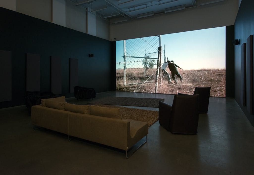 A large screen on a gallery wall in front of a sofa, rugs and chairs. A single-channel video is projected on the screen, depicting two people in a physical altercation by a chain link fence.