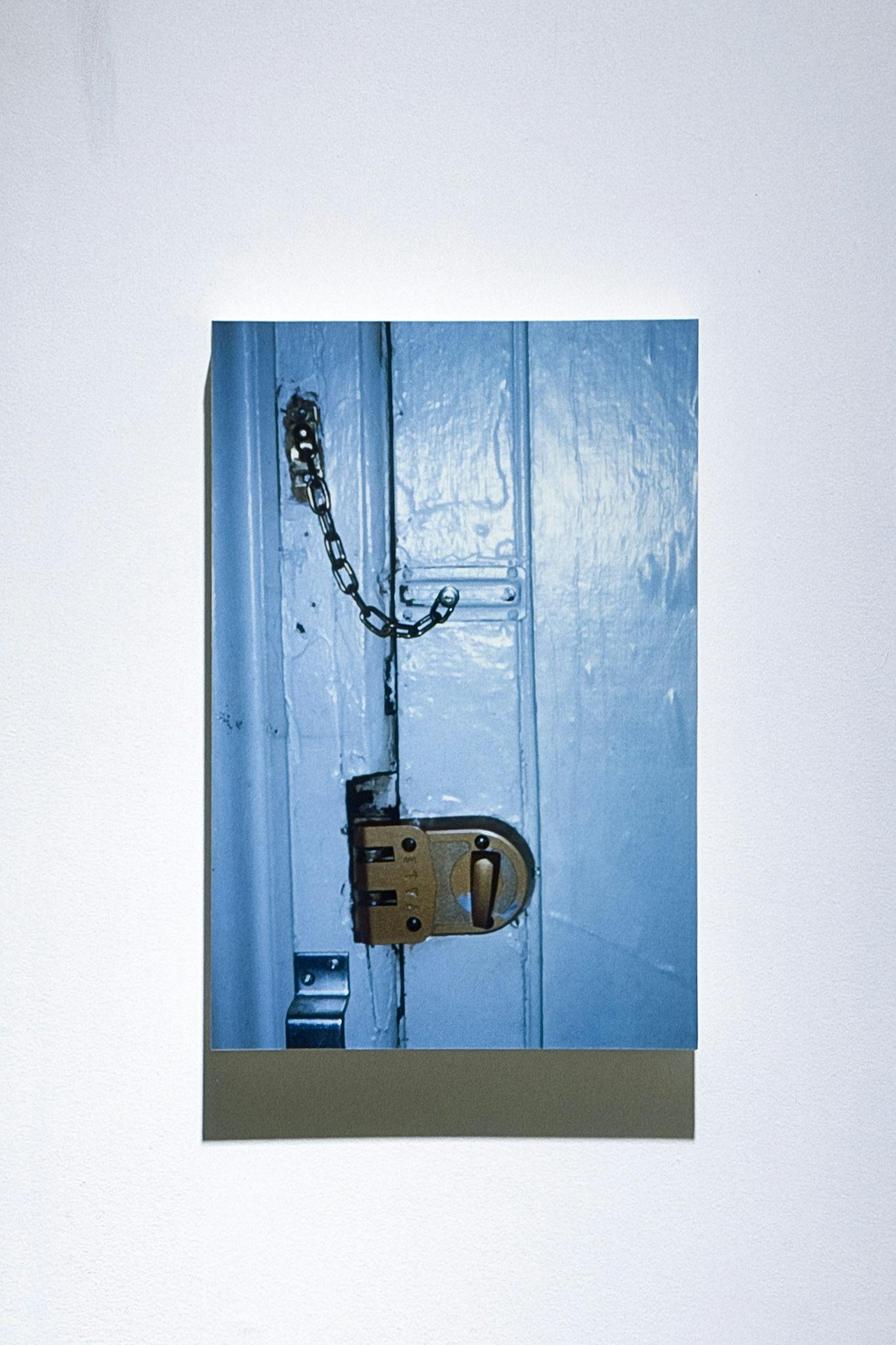 A rectangular-shaped photograph is mounted on the white gallery wall. This is a partial image of a blue door with a brown lock and a silver door chain.   