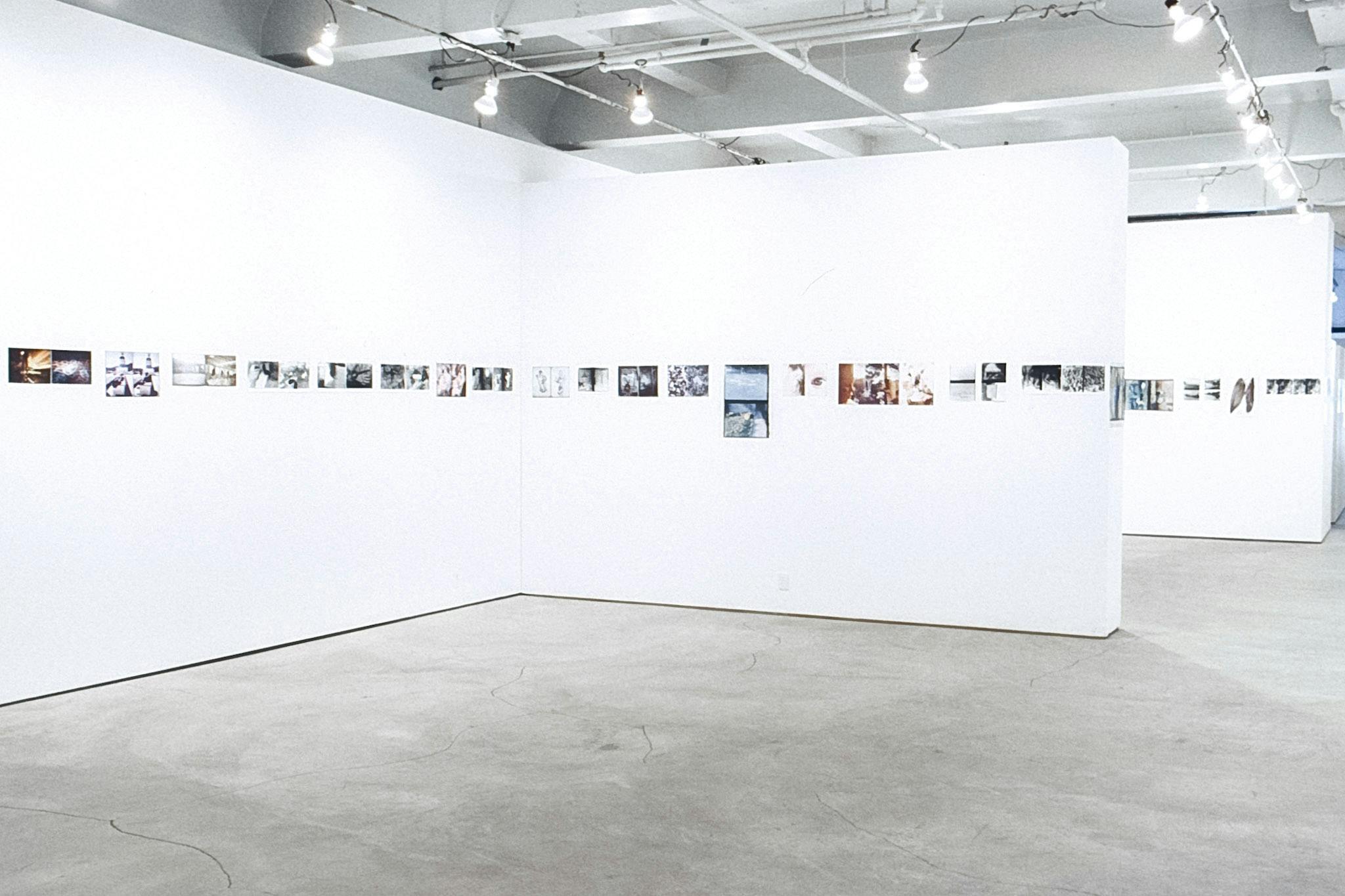 A gallery with a white wall devings the space. Several photos, both in colour and black and white, are arranged in a line across all walls. The photos vary in size and orientation. 