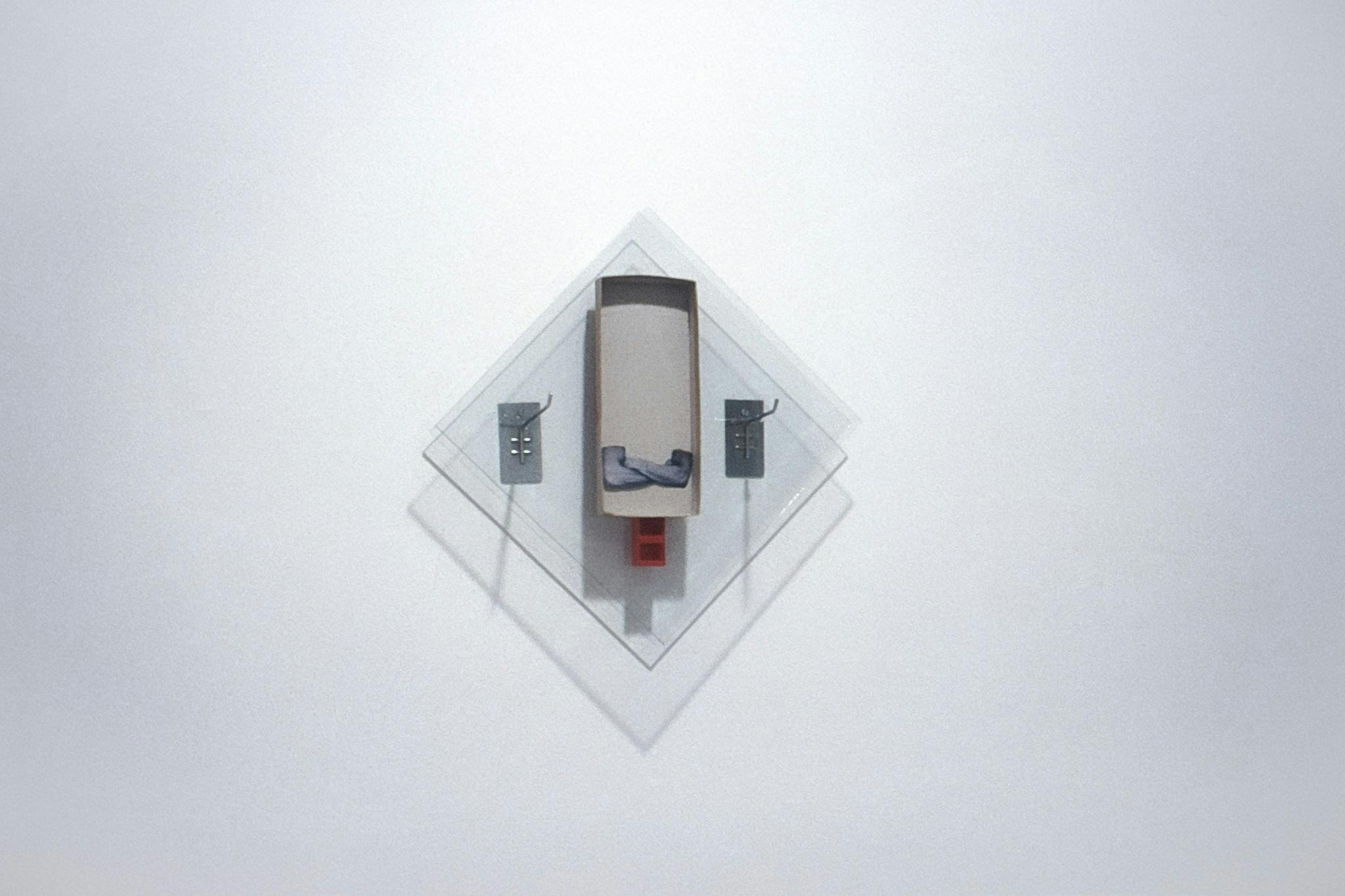 An artwork mounted on a white wall. The work is a piece of glass in a square diamond shape with a grey-brown rectangle and a red cylinder mounted on top. A photo of two crossed arms is pasted onto it. 