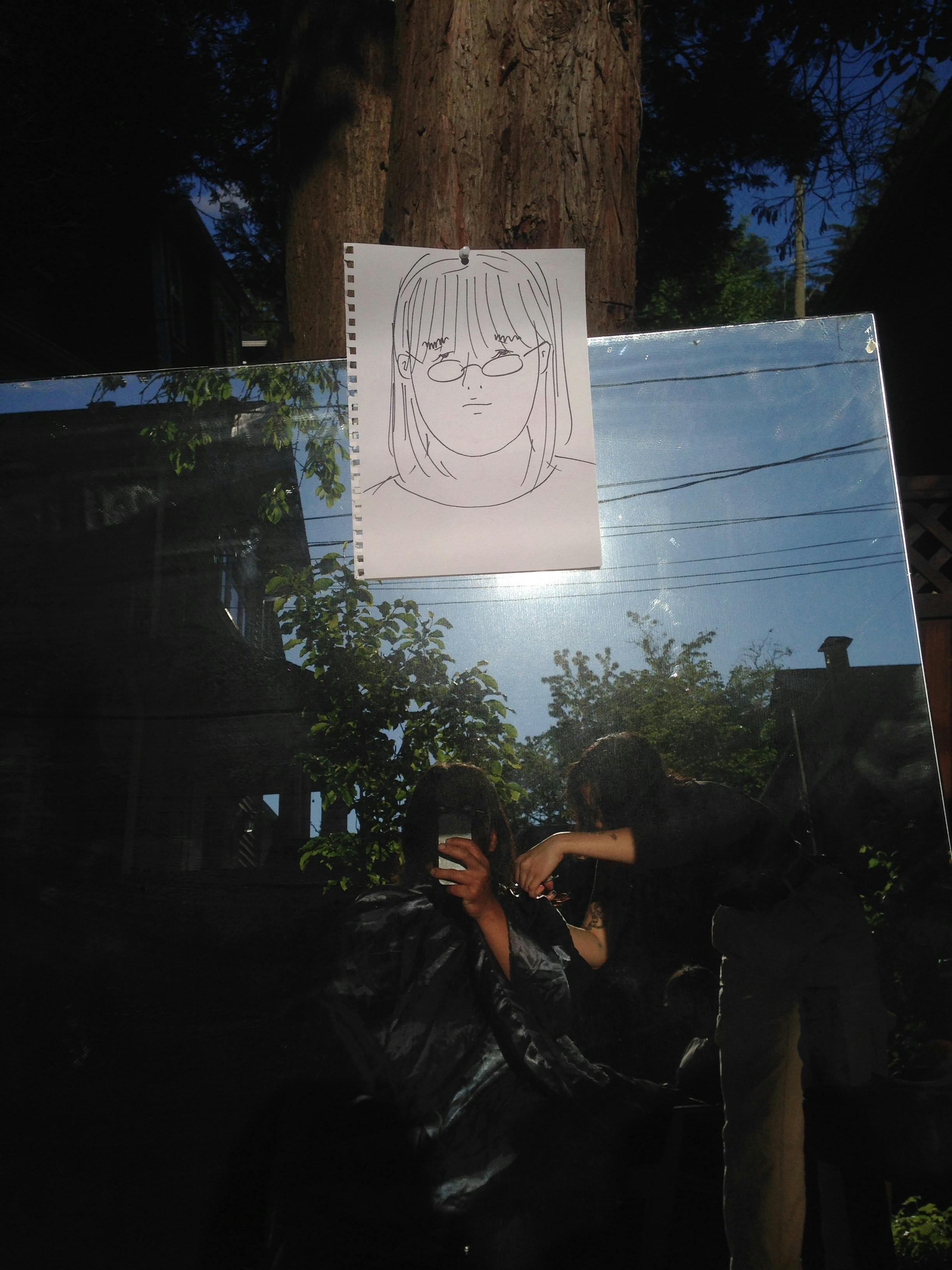 A photograph of a large mirror placed outside. A drawing of a person's face is attached to the top of the mirror. The reflection on the mirror shows people getting haircuts. 