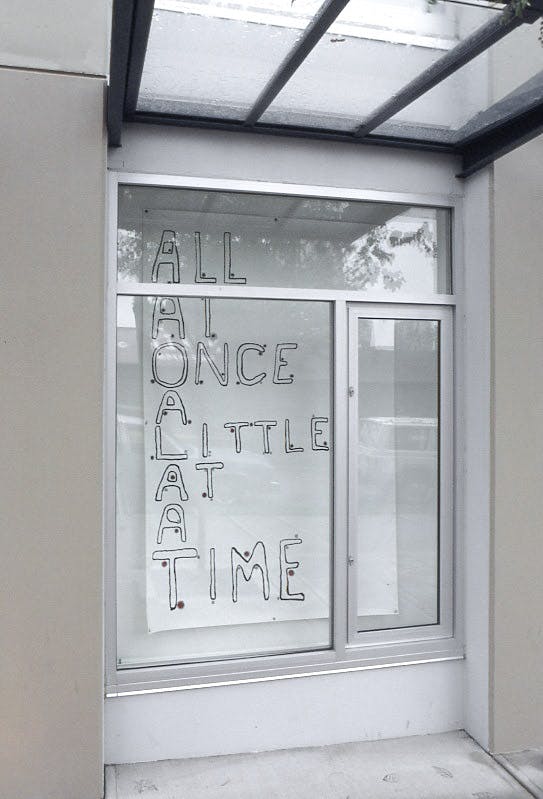 Jill Henderson’s text-based work is installed in one of CAG’s window spaces. The window in this image showcases a handwritten text that reads “all at once a little at a time”  in upper cases.