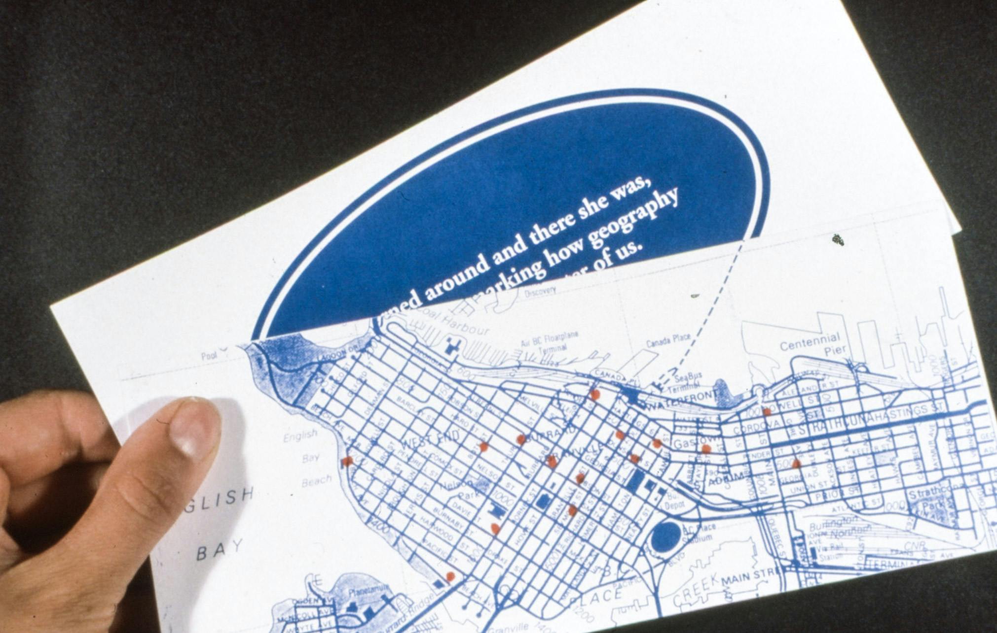 A closeup of a hand holding two horizontal papers. The one in front shows a map of downtown Vancouver and its neighbourhoods. The one in the back shows a blue oval with partially visible white text.